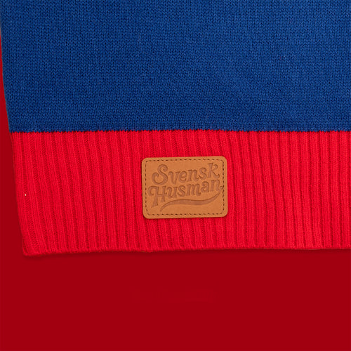 Blau och rot stickad jultröja med Schwedischhusman-logotyp på en läderlapp mot rot bakgrund.