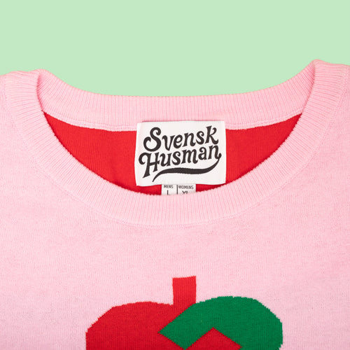 Rosa stickad jultröja med rött och grönt julhjärta och etikett från Svenskhusman på ljusgrön bakgrund.
