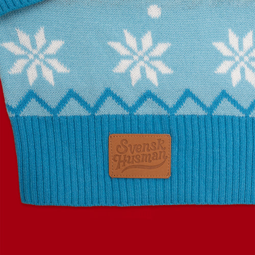 Blå stickad jultröja med vita snöflingor och brun läderlapp från Svenskhusman på röd bakgrund.